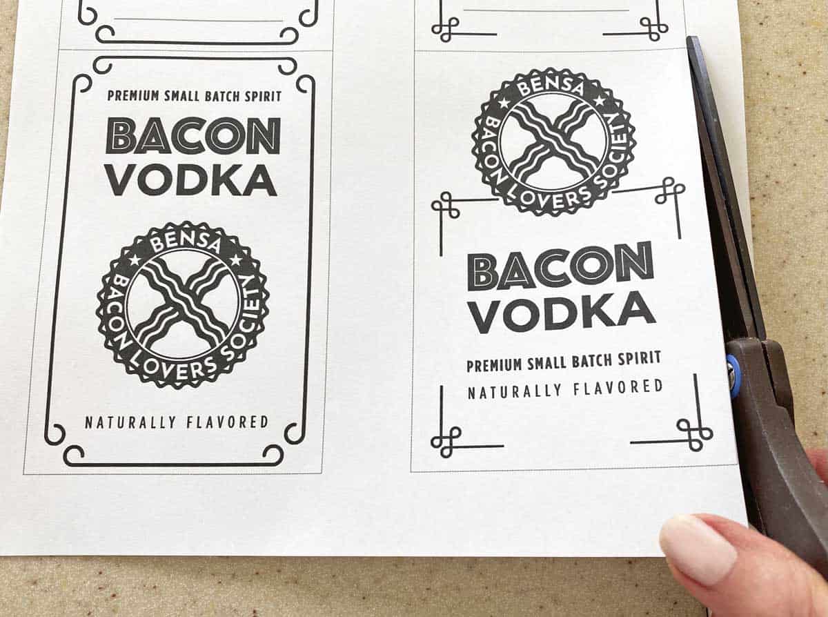 A hand show with a pair of scissors, cutting a sheet of bacon vodka labels